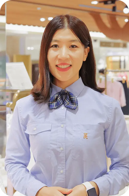 薄紫色のシャツにリボンタイを着用し、笑顔で立つ女性ファッションアドバイザーL.W.さんのポートレート