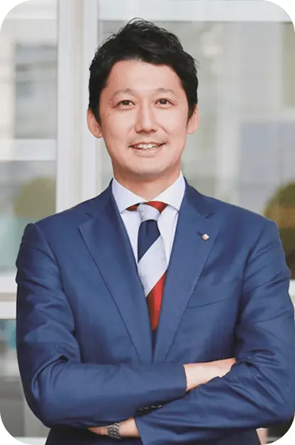 紺色のスーツに赤いネクタイを着用し、笑顔で腕を組むS.Nさんのポートレート