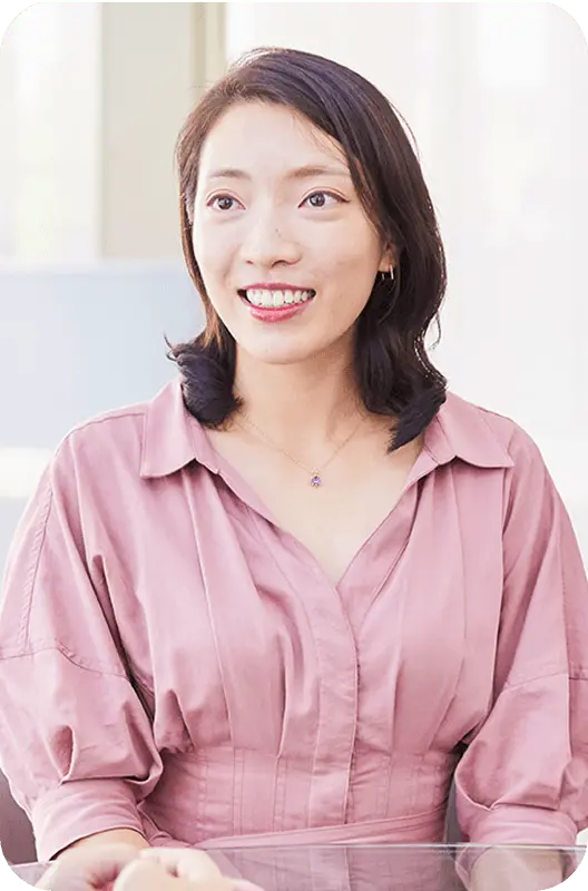 ピンク色のシャツを着て笑顔で立つ女性社員Y.Jさんのポートレート